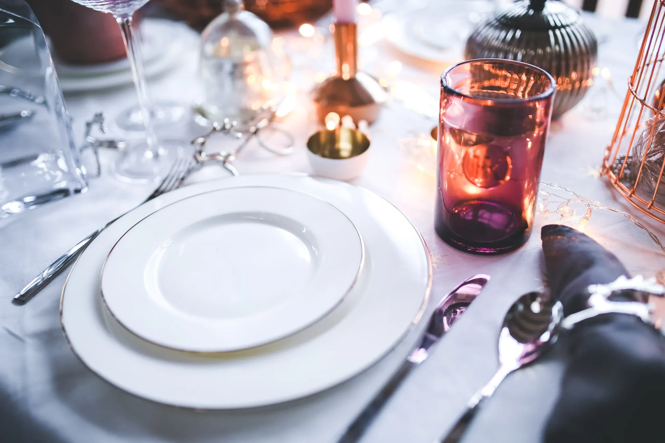 A dinner table set with white plates, silver cutlery and glassware glowing in warm candlelight