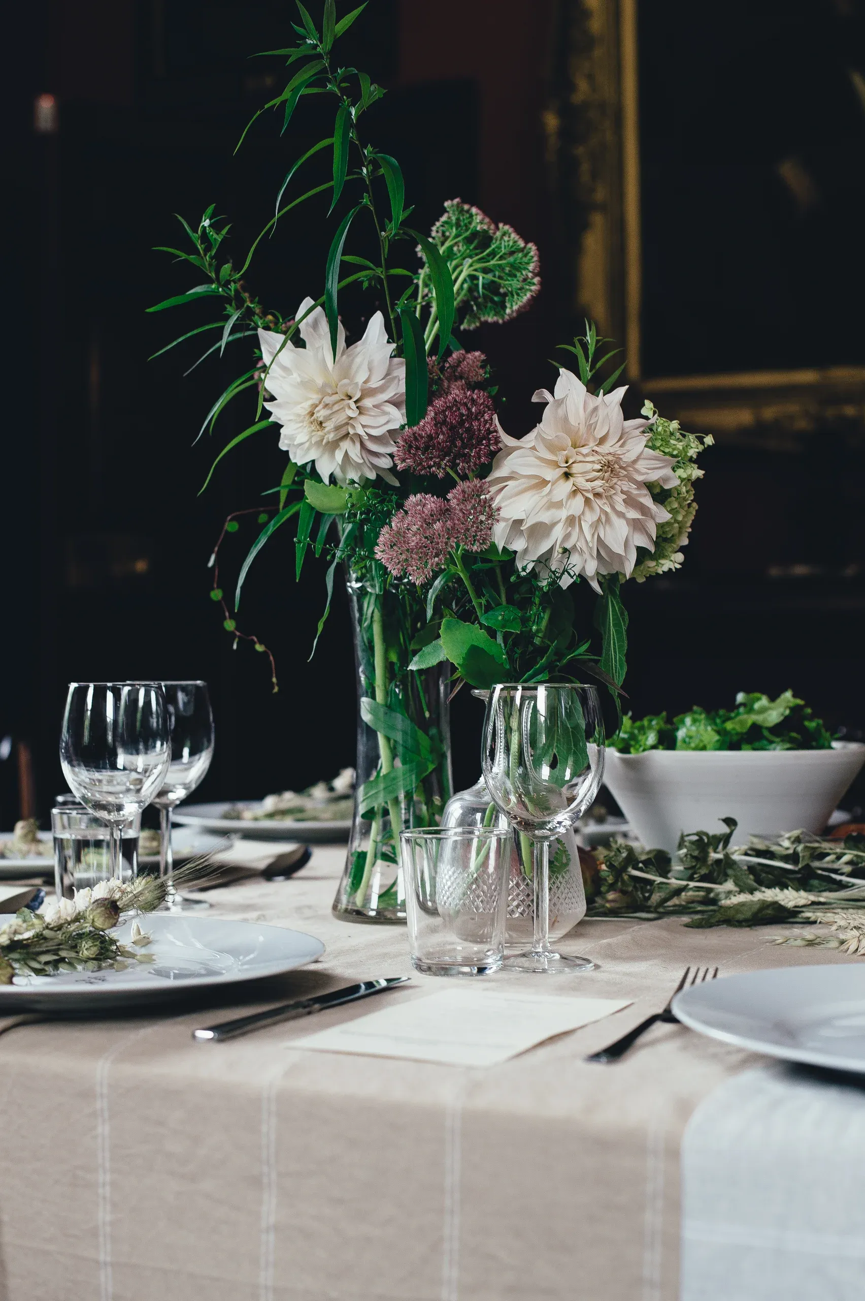 Pale dahlias and greenery in a glass vase on a linen dressed table in low light