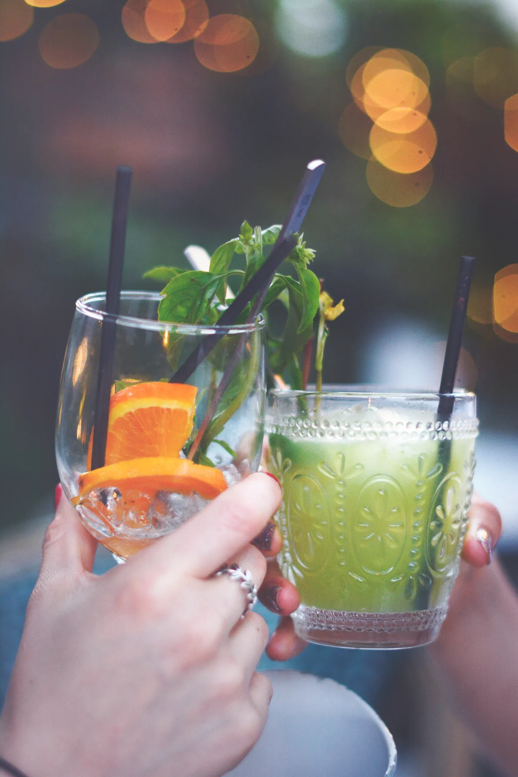 Two hands raising cocktails with citrus and mint in a toast, with warm lights blurred behind